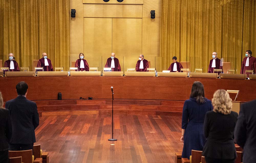 Euroopan unionin tuomioistuimessa tuomarit seisovat kaarevan pöydän takana punaisissa kaavuissa ja maskit kasvoilla. 